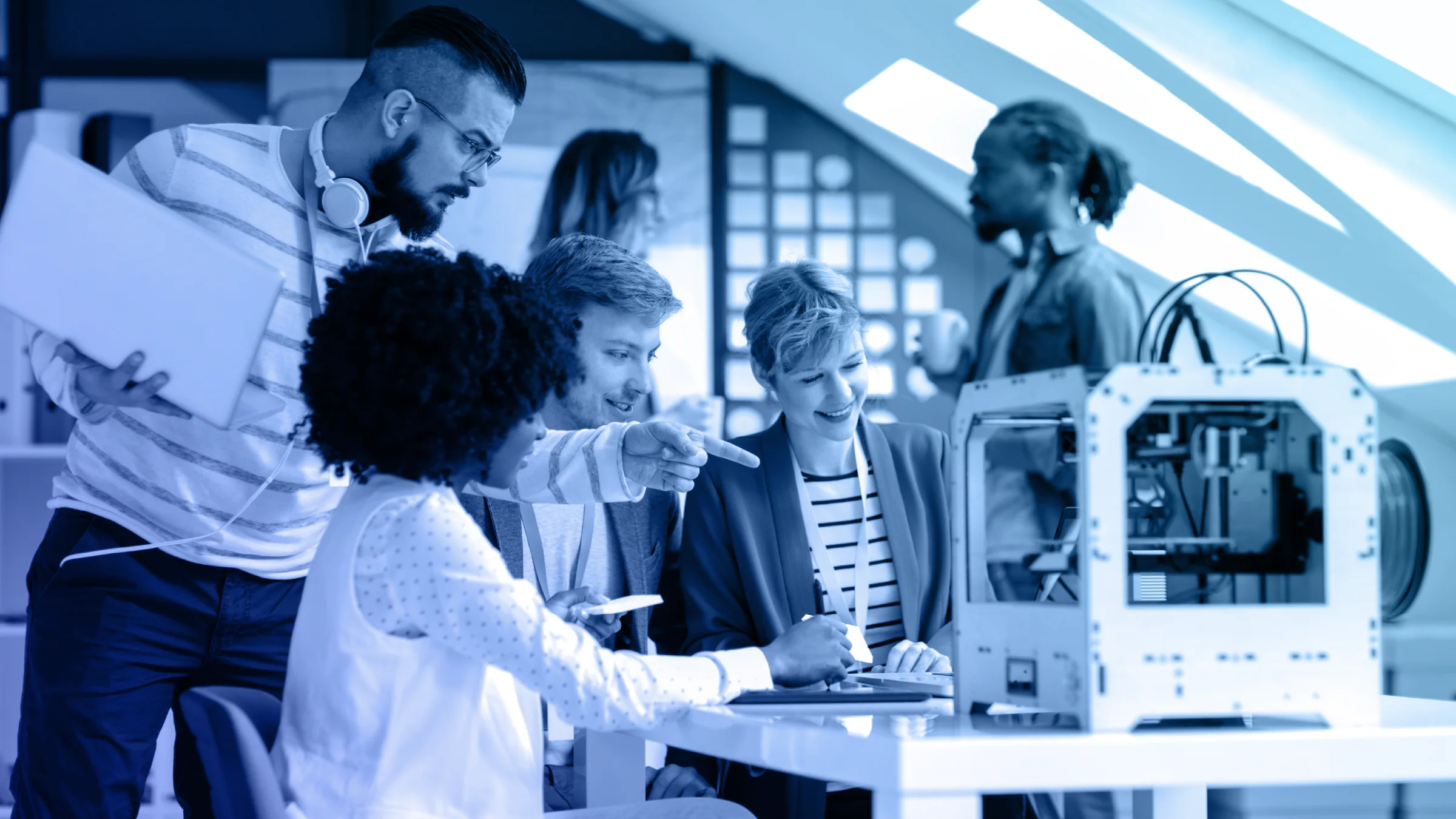A group of diverse professionals collaborating around a laptop in a modern workspace, with a 3D printer on the table. The image is tinted in blue tones, emphasizing the technology and teamwork in an innovative setting.